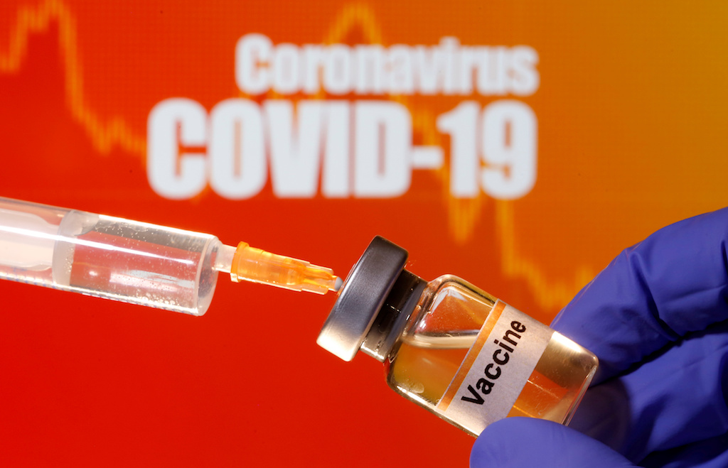 A small bottle labeled with a "Vaccine" sticker is held near a medical syringe in front of displayed "Coronavirus COVID-19" words in this illustration.