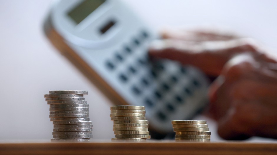File: A hand holds a calculator behind three stacks of coins.