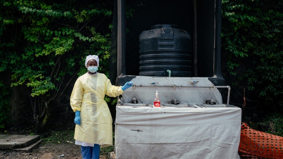 A health worker, wearing single-use protective clothing and a surgical cap, stands at a checkpoint where she oversees hand washing and temperature screening. AFP/Jospin Mwisha