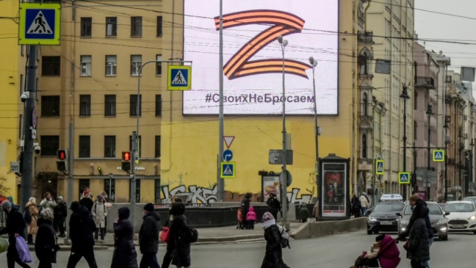 Pedestrians cross a street in front of a billboard displaying the symbol "Z" in the colours of the ribbon of Saint George and a slogan reading: 'We don't give up on our people'