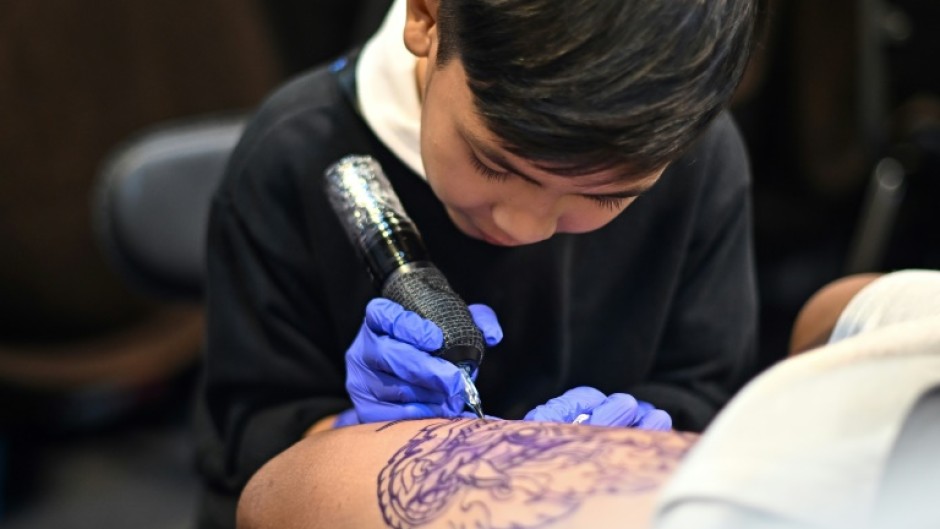 Wielding a cumbersome tattoo gun, nine-year-old Napat Mitmakorn inks the pattern of a fanged serpent on a man's upper thigh