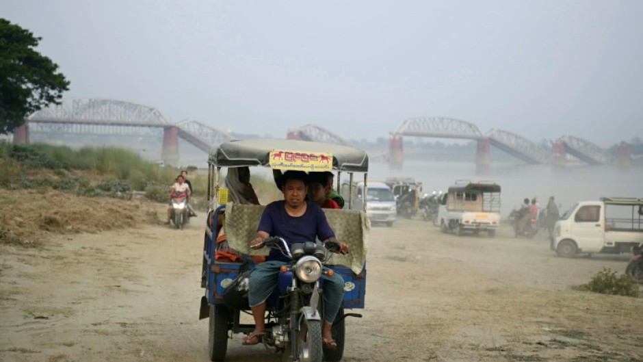 A tuk tuk driver ferries passengers alongg the bank of the Irrawaddy River near Sagaing, with the collapsed Ava Bridge in the background