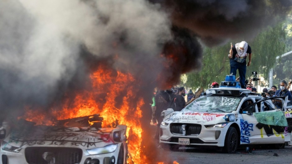 Demonstrators smash the windshield of a vehicle next to a burning self-drive vehicle as protesters clash with law enforcement in Los Angeles, California, on June 8, 2025