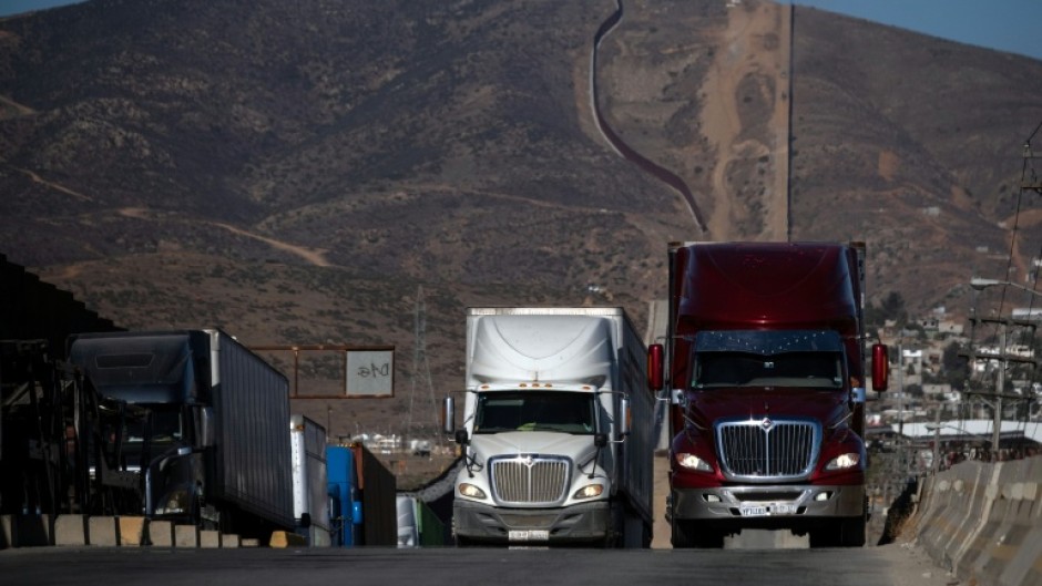 Cargo trucks queue next to the border wall before crossing into Tijuana, Mexico
