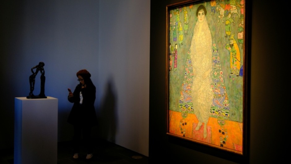 The piece represents the daughter of Klimt's main patron dressed in a white imperial Chinese dress, standing before a blue tapestry with Asian-inspired motifs