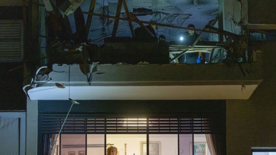 A woman stands at a window as first responders and security forces inspect a damaged apartment above after the building was hit by an Iranian missile strike in Ramat Gan, on the outskirts of Tel Aviv, early on March 18, 2026