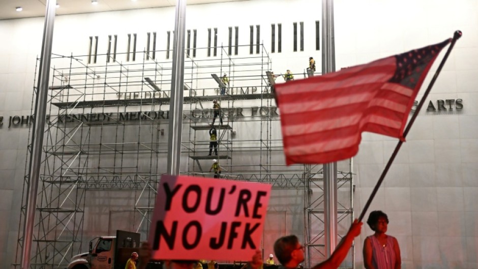 Protesters wave a US flag and hold a sign reading 'You're no JFK' as workers build scaffolding at the Kennedy Center ahead of the removal of Donald Trump's name