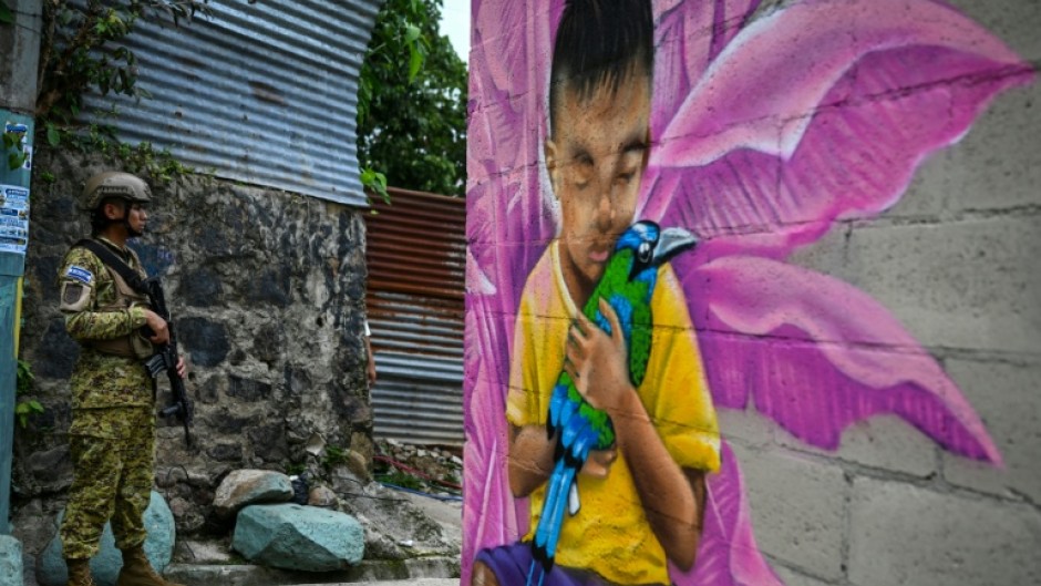A Salvadoran soldier stands guard in a narrow passage in the 10 de Octubre community, in front of a mural painted by artist Carlos Clímaco, in San Marcos, El Salvador, on October 2, 2026. The artistic project aims to transform the identity of a community historically affected by gang violence.