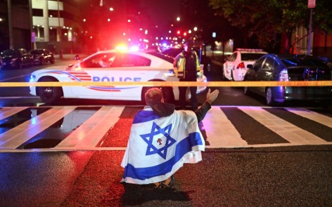A man draped in the Israeli flag, bearing a cross and the name 'Jesus' at its center, gestures as Metropolitan Police officers secure the area outside the Capital Jewish Museum
