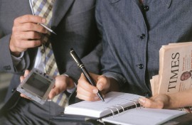 Close-up of hands of woman in suit writing on diary, hands of man in suit holding a palm-pilot at her side.