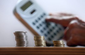 File: A hand holds a calculator behind three stacks of coins.