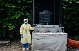 A health worker, wearing single-use protective clothing and a surgical cap, stands at a checkpoint where she oversees hand washing and temperature screening. AFP/Jospin Mwisha