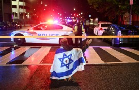A man draped in the Israeli flag, bearing a cross and the name 'Jesus' at its center, gestures as Metropolitan Police officers secure the area outside the Capital Jewish Museum