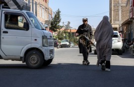 An Afghan burqa-clad woman walks past a Taliban security personnel standing guard outside a mosque in Shahrak-e-Almahdi, Jebrail district of Herat Province on June 11, 2026