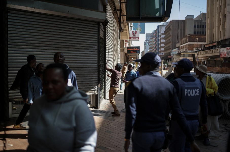 South African Police Service (SAPS) officers watch as a worker closes the shutters of a shop while protestors pass by during a protest march against undocumented migrants organised by “March and March” in Johannesburg on April 29, 2026. 