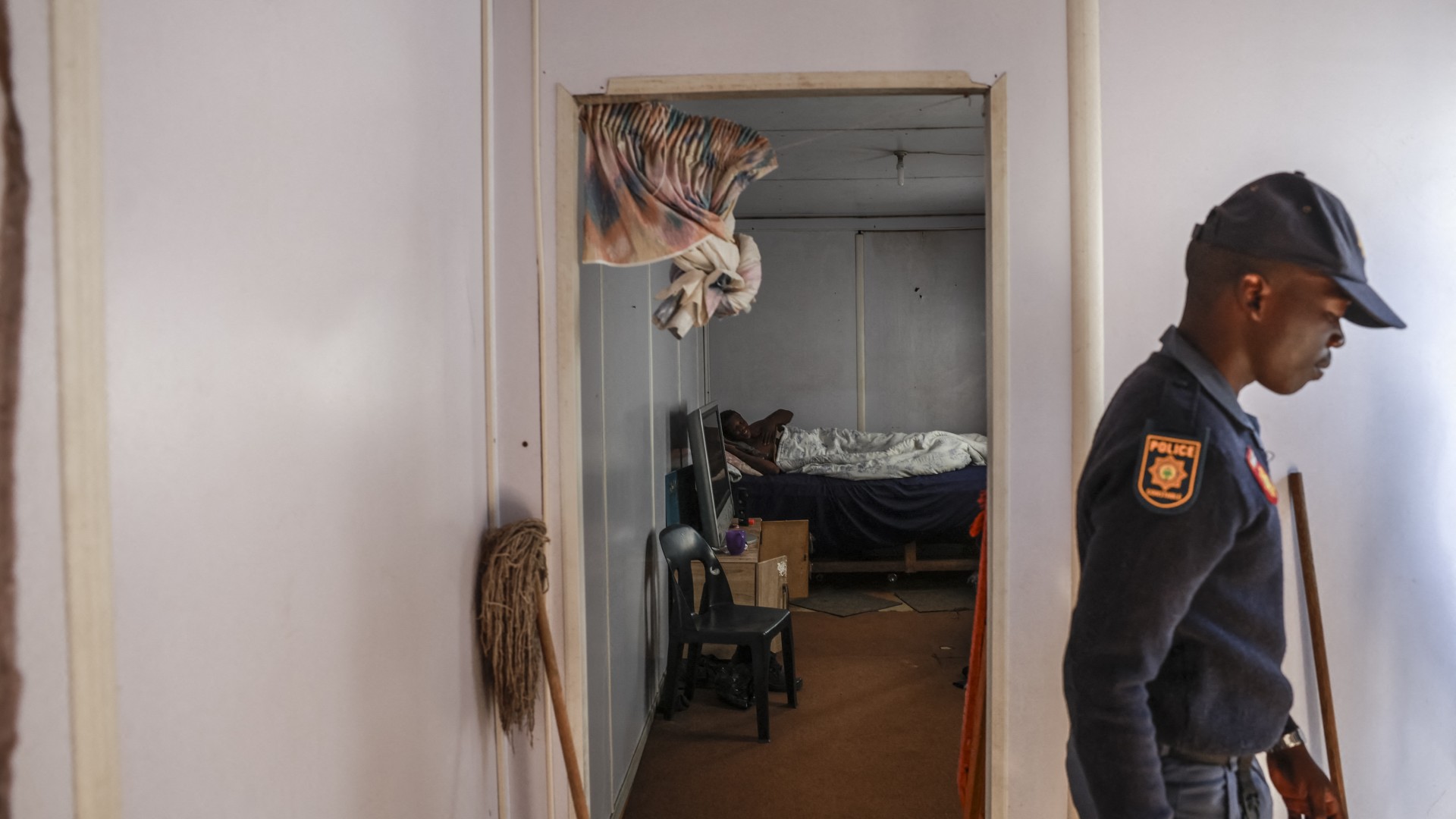 A hostel dweller lies on a bed while South African Police Service (SAPS) officer leaves after raiding his home to retrieve looted goods during a protest at Diepkloof Hostel in Soweto on May 19, 2025.