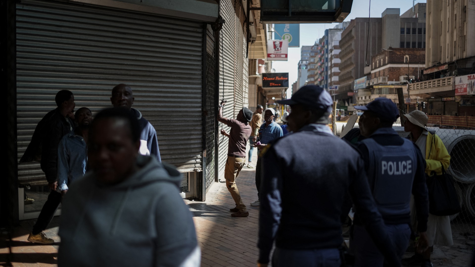 South African Police Service (SAPS) officers watch as a worker closes the shutters of a shop while protestors pass by during a protest march against undocumented migrants organised by “March and March” in Johannesburg on April 29, 2026.