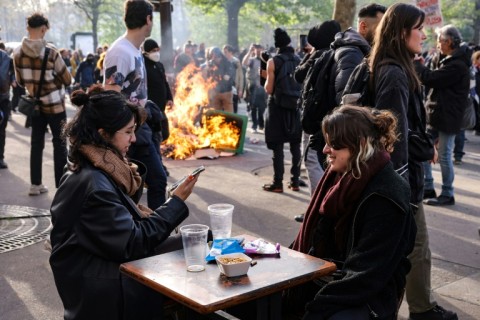 Two girls have a drink at a cafe terrace as a garbage can is set ablaze nearby during a demonstration on the 11th day of action after the government pushed a pensions reform through parliament without a vote
