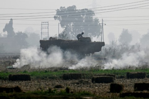 A worker loads a tractor with paddy straw bales as smoke rises from smoldering leftover stubble