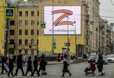 Pedestrians cross a street in front of a billboard displaying the symbol "Z" in the colours of the ribbon of Saint George and a slogan reading: 'We don't give up on our people'