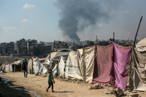 People walk past makesfift shelters as smoke billows east of Gaza City, following Israeli bombardment