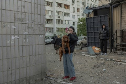 A woman holding her dog and takes photos of the residential building partially destroyed