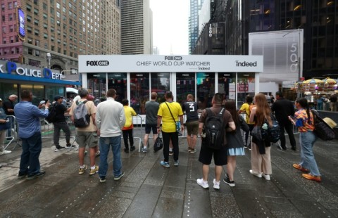 People look on outside a cube-like studio in New York's Times Square, where two men are paid by broadcaster Fox to watch all 104 World Cup matches