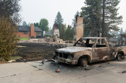 A burned pickup truck sits in the driveway of a burned residential home with an untouched home in the background in the Balboa neighborhood of Spokane, Washington, on August 2, 2026.