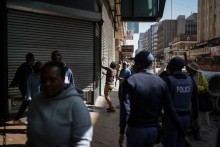 South African Police Service (SAPS) officers watch as a worker closes the shutters of a shop while protestors pass by during a protest march against undocumented migrants organised by “March and March” in Johannesburg on April 29, 2026.