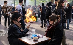 Two girls have a drink at a cafe terrace as a garbage can is set ablaze nearby during a demonstration on the 11th day of action after the government pushed a pensions reform through parliament without a vote