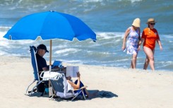 Beachgoers walk along the water as US President Joe Biden and US First Lady Jill Biden sit under an umbrella in Rehoboth Beach, Delaware