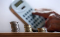 File: A hand holds a calculator behind three stacks of coins.