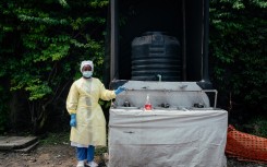 A health worker, wearing single-use protective clothing and a surgical cap, stands at a checkpoint where she oversees hand washing and temperature screening. AFP/Jospin Mwisha