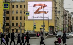 Pedestrians cross a street in front of a billboard displaying the symbol "Z" in the colours of the ribbon of Saint George and a slogan reading: 'We don't give up on our people'