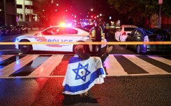 A man draped in the Israeli flag, bearing a cross and the name "Jesus" at its center, gestures as Metropolitan Police officers secure the area outside the Capital Jewish Museum