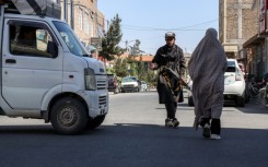 An Afghan burqa-clad woman walks past a Taliban security personnel standing guard outside a mosque in Shahrak-e-Almahdi, Jebrail district of Herat Province on June 11, 2026