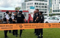 Damaged windows can be seen in the distance as police gather near the scene of a shooting in Montreal, which left three people dead