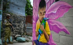 A Salvadoran soldier stands guard in a narrow passage in the 10 de Octubre community, in front of a mural painted by artist Carlos Clímaco, in San Marcos, El Salvador, on October 2, 2026. The artistic project aims to transform the identity of a community historically affected by gang violence.